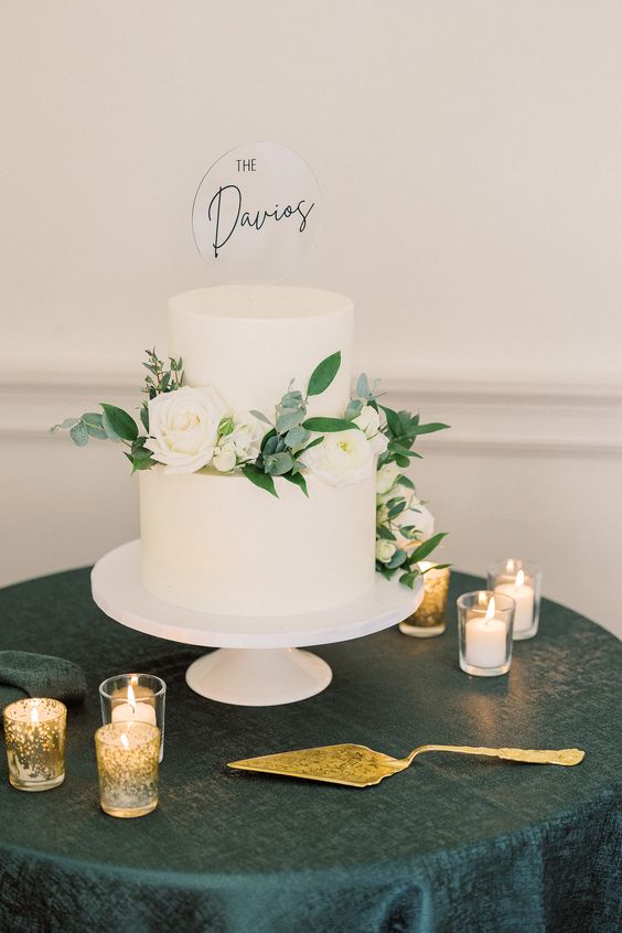 a white buttercream wedding cake decorated with white blooms and greenery, with a clear acrylic cake topper with black calligraphy