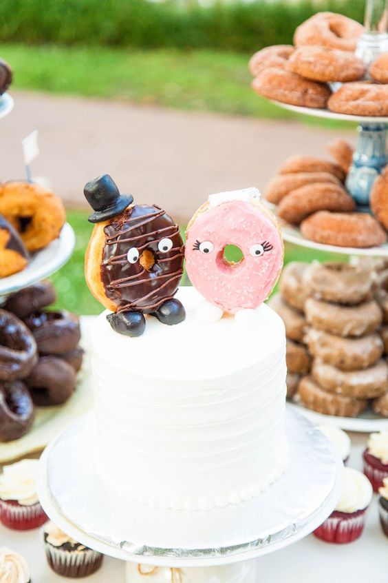 a white buttercream wedding cake topped with glazed donuts dressed up like a marrying couple look very cute and nice
