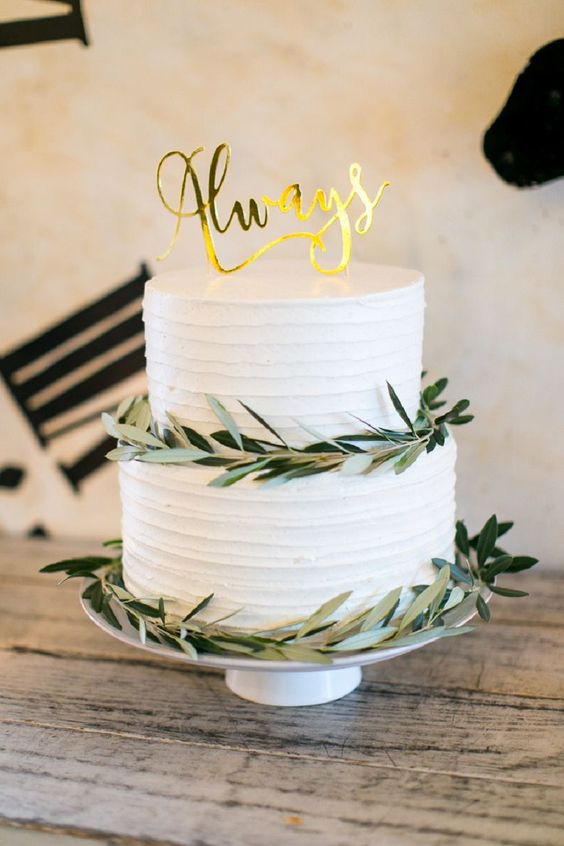 a white buttercream wedding cake topped with greenery, with a gold calligraphy cake topper is a cool idea for a nerdy wedding