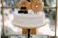 a white buttercream wedding cake topped with marshmallows, glazed donut cake toppers dressed up like a marrying couple
