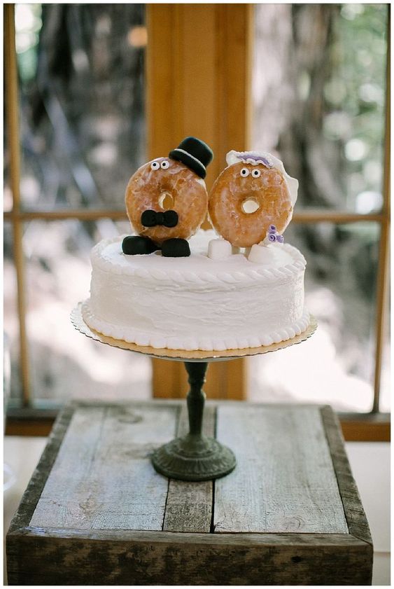 a white buttercream wedding cake topped with marshmallows, glazed donut cake toppers dressed up like a marrying couple