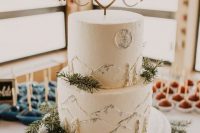 a white buttercream wedding cake with painted mountains, a moon and trees, with evergreens and a monogram and heart cake topper