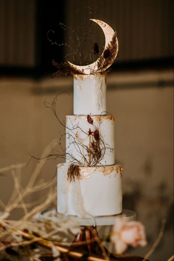 a white celestial wedding cake with gold foil, dried blooms, a moon and some twigs on top is a chic and lovely idea