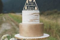 a woodland wedding cake with a white and a gold tier, with a mountain and calligraphy topper is a catchy and bold idea to try