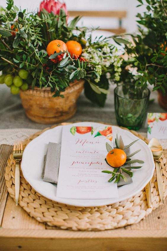 a Mediterranean summer wedding tablescape with a woven charger, potted greenery with fruits and gold cutlery