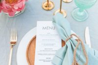 a bright beach table with a light blue tablecloth, turquoise candles, aqua green glasses, a blue napkin, a wooden plate and pink blooms