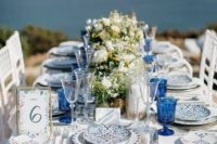 a bright blue beach table with bold glasses, printed plates, neutral floral and greenery centerpieces and a pretty table number
