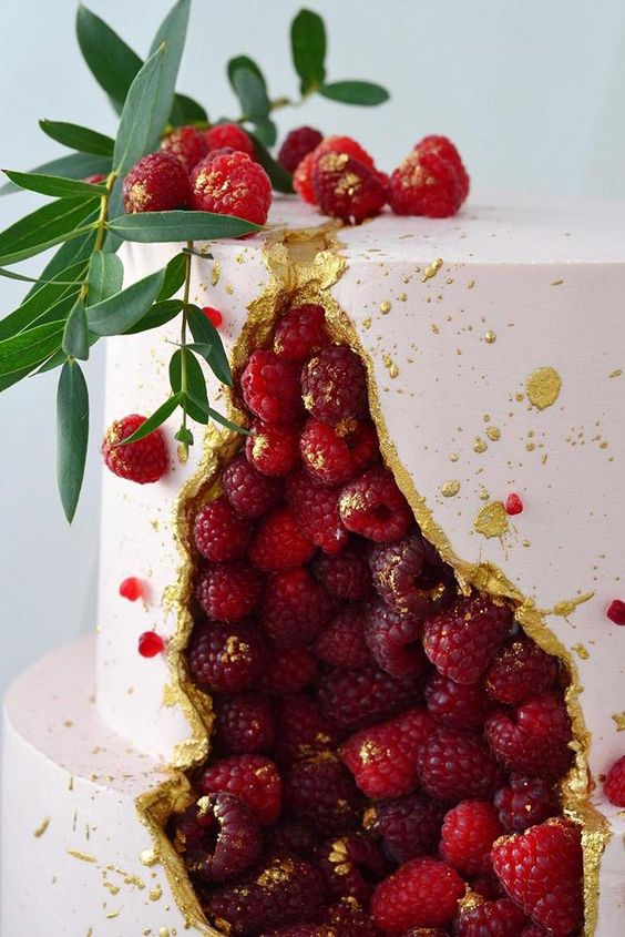 a pink broken wedding cake with gold edges and gilded raspberries plus greenery on top and gold splatters is wow