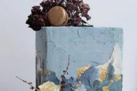 a rectangular wedding cake with textured blue buttercream and gold accents, with dried deep purple blooms on top and some gilded macarons for a modern wedding