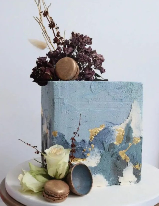 a rectangular wedding cake with textured blue buttercream and gold accents, with dried deep purple blooms on top and some gilded macarons for a modern wedding