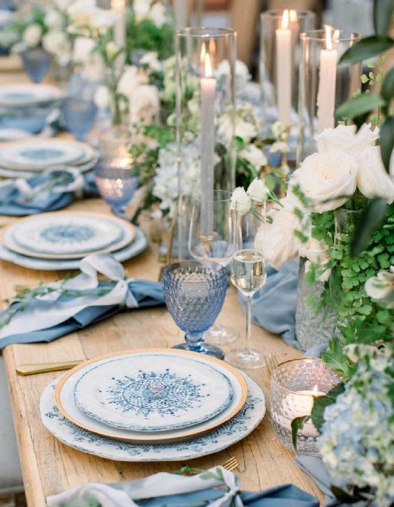 a refined summer wedding table in blues, with printed plates, white roses, greenery, blue candles and glasses