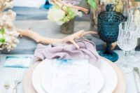 a seaside wedding tablescape with a dusty pink napkin, a blue runner, watercolor cards, deep blue glasses, white floral centerpieces and driftwood