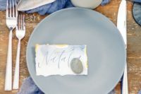 an oceanside wedding tablescape with a deep blue runner and napkins, grey porcelain, candles, seashells, watercolor cards
