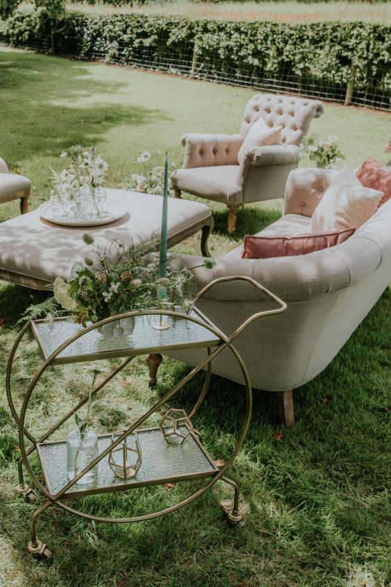 a beautiful and exquisite neutral wedding lounge with elegant seating furniture, a glass and brass bar cart, blooms and greenery and a green candle