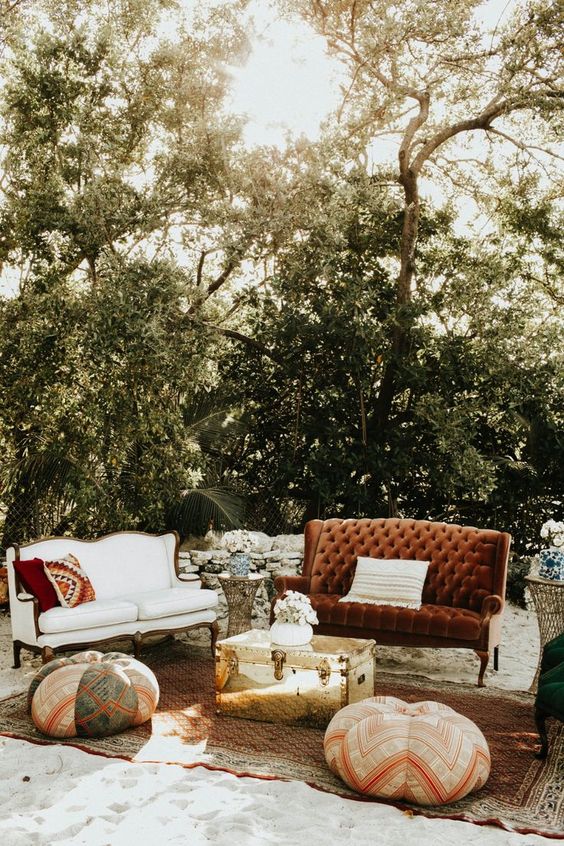 a bold boho lounge with a white and a rust-colored sofa, printed pillows, a gold chest for storage, printed poufs and a rug