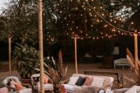 a bold boho wedding lounge with string lights, white rattan furniture, coffee tables, leather poufs, pampas grass and candle lanterns