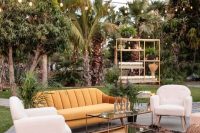 a bold mid-century modern outdoor wedding lounge with a yellow velvet sofa, neutral chairs, a printed rug and a glass table, poufs