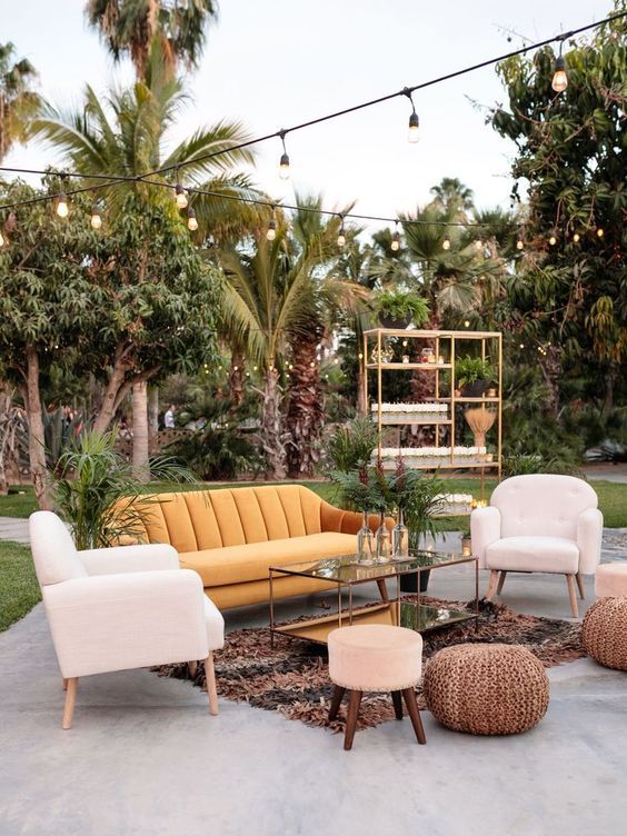 a bold mid-century modern outdoor wedding lounge with a yellow velvet sofa, neutral chairs, a printed rug and a glass table, poufs