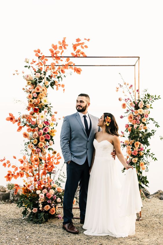 a bold modern wedding arch covered with blush, light pink, orange and yellow blooms and greenery is wow