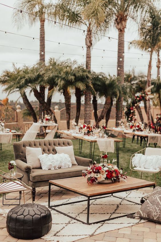 a bold outdoor wedding lounge with a grey loveseat, metal chairs, poufs and pillows, a rug, a low coffee table and bold blooms