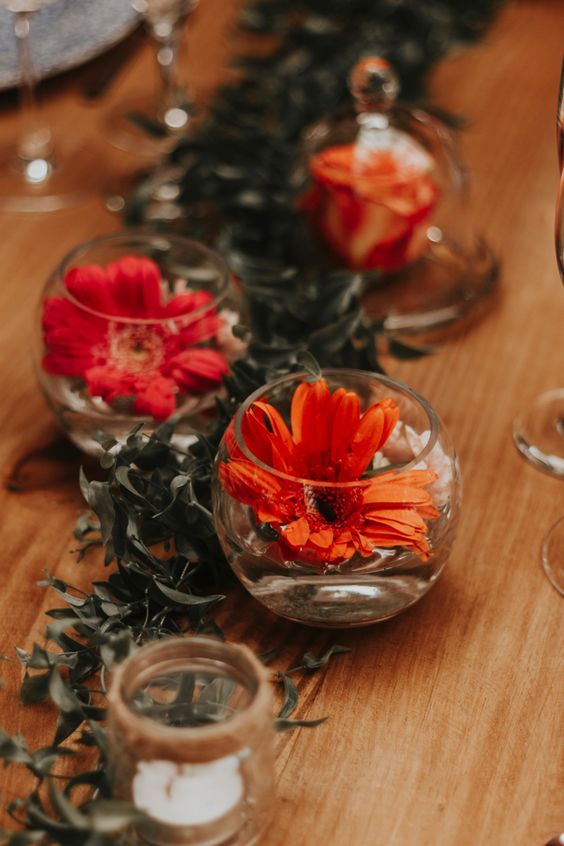 a bold wedding table runner of greenery, orange and red blooms in glass bowls and candles is gorgeous