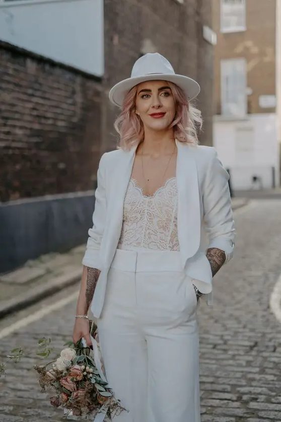 a bride wearing a white pantsuit with a cropped blazer, a white lace bodice and a white fedora hat plus pink hair looks fantastic