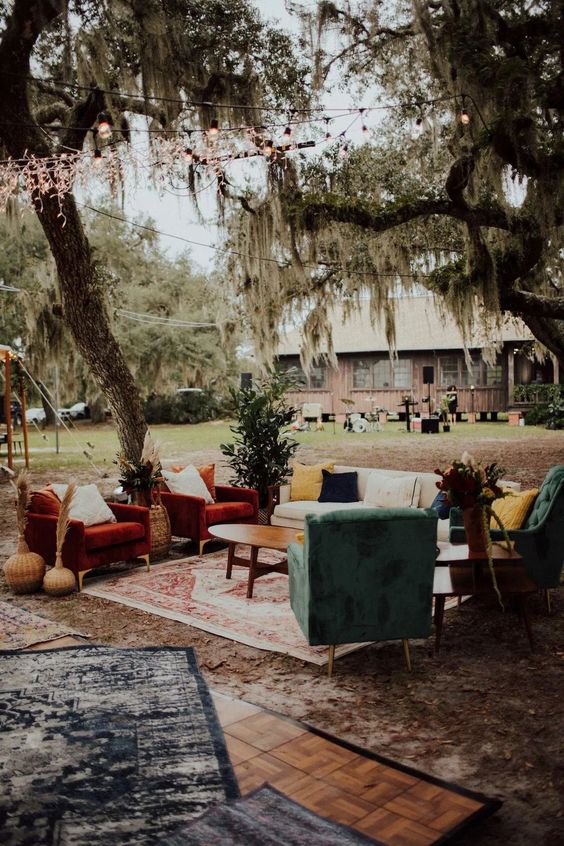 a bright boho outdoor wedding lounge with red and green chairs, a neutral sofa and bright pillows, a coffee table and layered rugs