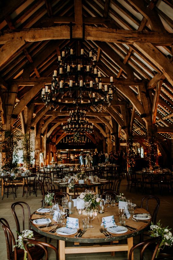 A cozy barn wedding reception space with large candle chandeliers, greenery and neutral blooms and wooden uncovered tables