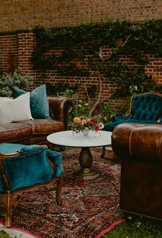 a dark and moody boho wedding lounge with teal vintage chairs, leather sofas with blue pillows, a printed rug and a round table