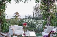 a garden wedding lounge with white seating furniture, a coffee table, purple blankets and pink blooms, a printed rug for more coziness