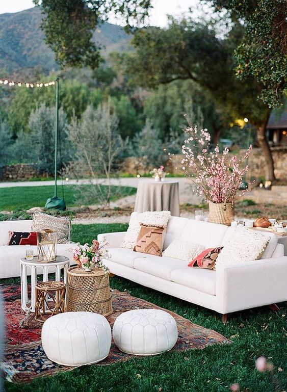 a lovely boho outdoor wedding lounge with white seating furniture, printed pillows, an arrangement of coffee tables, white leather poufs and some blooming branches