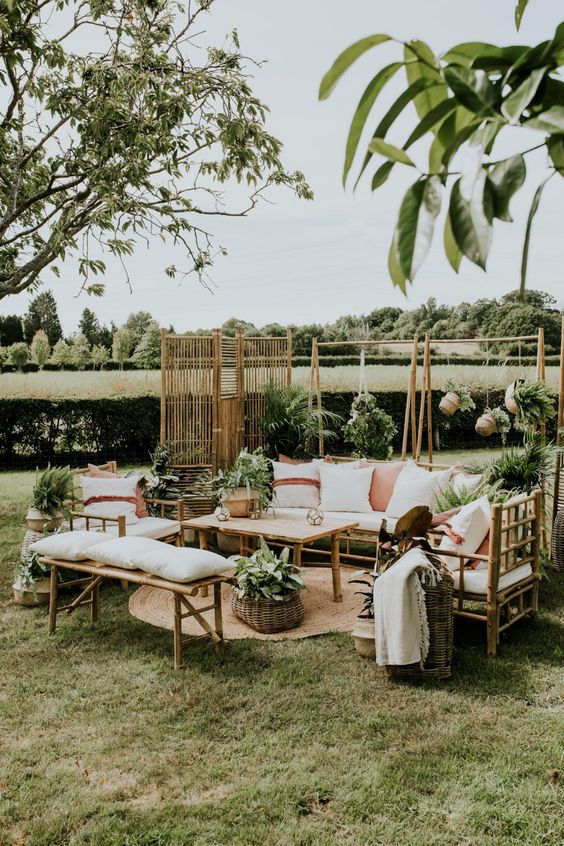 a lovely outdoor wedding lounge with bamboo furniture with neutral upholstery, neutral and pink pillows, potted greenery all around