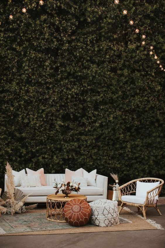 a neutral boho glam wedding lounge with a white leather sofa, a neutral rattan chair, neutral and pink pillows, a coffee table and some poufs plus pampas grass