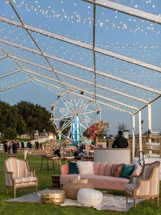 a pastel outdoor wedding lounge with a pink sofa and colorful pillows, striped chairs, poufs, layered rugs and lights over it