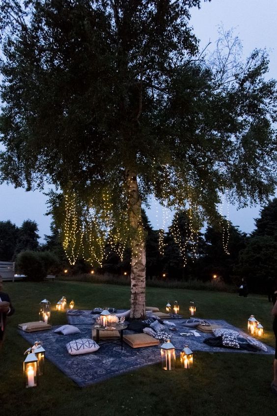 a relaxed and cool outdoor wedding lounge with rugs and pillows on the grass, candle lanterns and lights hanging on the tree