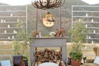 a rustic outdoor wedding lounge with a leather sofa, earthy-tone chairs, a round table, a printed rug, a faux fireplace with firewood and an antler chandelier