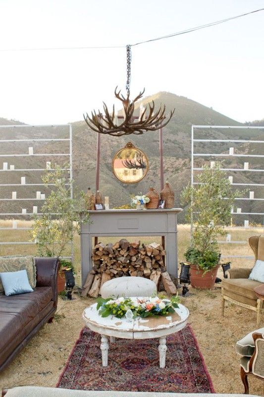 a rustic outdoor wedding lounge with a leather sofa, earthy-tone chairs, a round table, a printed rug, a faux fireplace with firewood and an antler chandelier