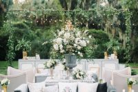 a sophisticated outdoor wedding lounge with dusty blue sofas and neutral chairs, glass coffee tables and white blooms and greenery