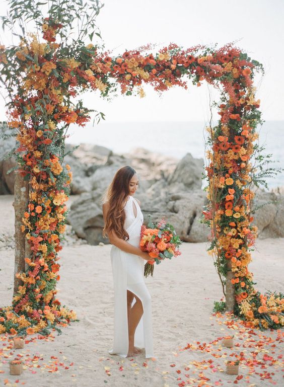 a super bold wedding arch covered with orange, red and rust blooms and lots of greenery is a gorgeous idea for a summer or fall wedding arch