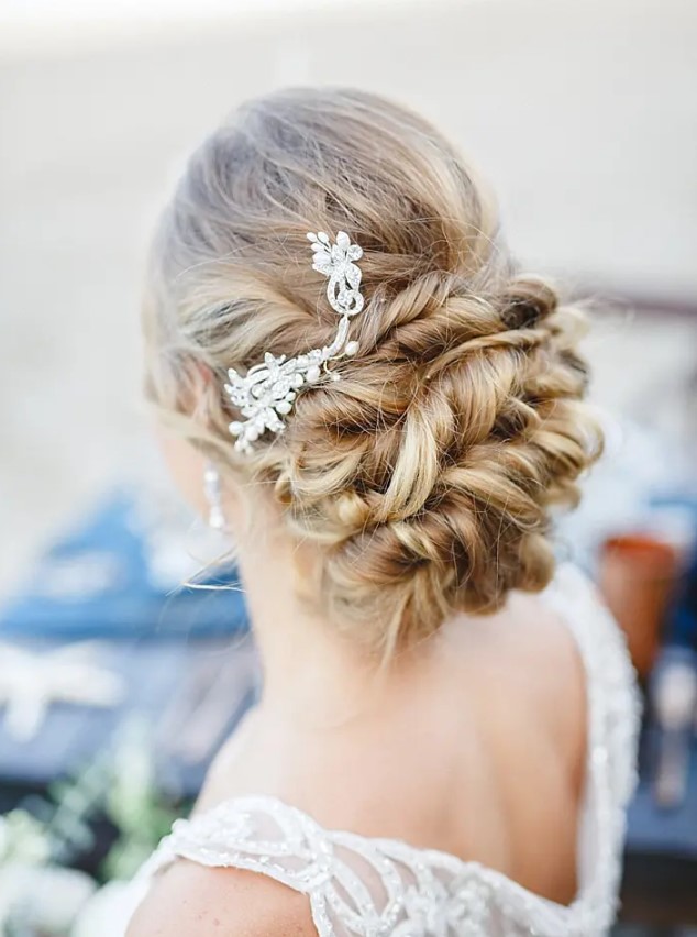 a twisted low updo on long hair with a rhinestone hairpiece on one side for a refined look at a beach or some other wedding