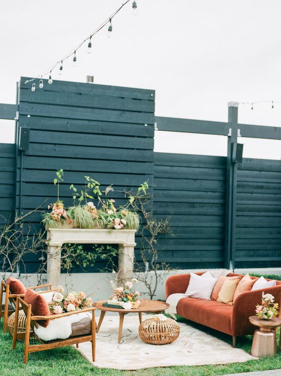 a welcoming outdoor wedding lounge with a faux fireplace, a rust-colored sofa with neutral pillows, taupe chairs with rust pillows and coffee tables