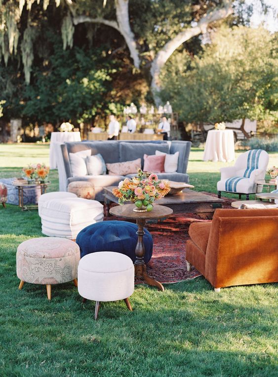 an eclectic boho wedding lounge with a grey sofa and a rust-colored seat, a couple of coffee tables, some poufs and bright blooms