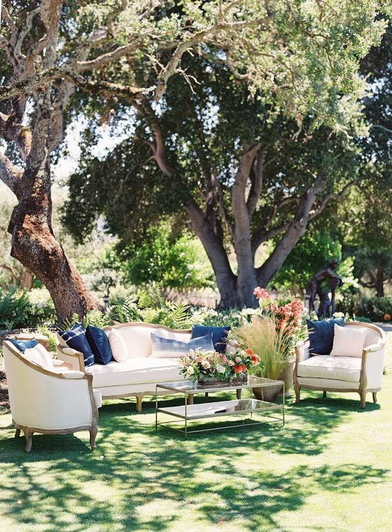 an elegant and refined outdoor wedding lounge with neutral furniture, a glass coffee table and bold blooms and greenery
