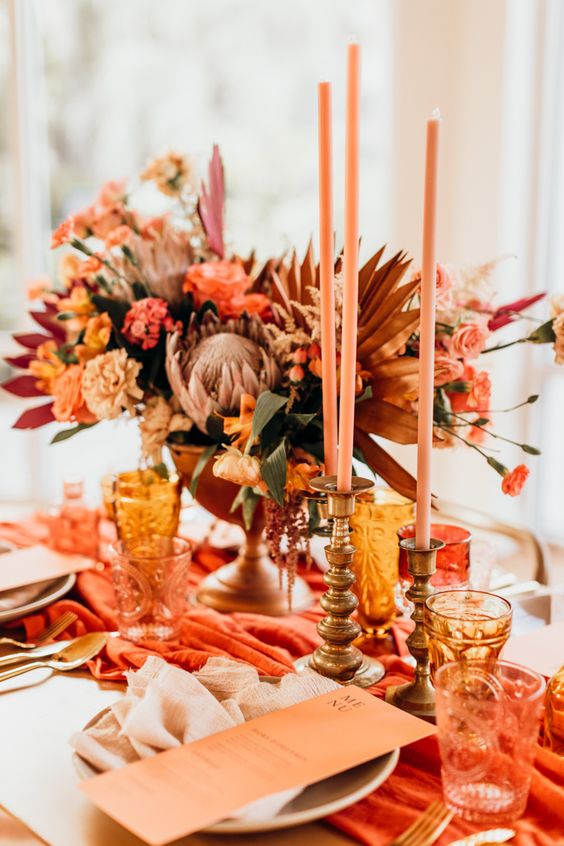 an extra bold orange and red wedding tablescape with a rust colored table runner, orange menus and candles, orange, peachy and red blooms and leaves