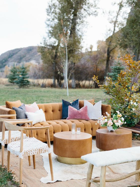 an outdoor fall wedding lounge with a mustard sofa, wooden chairs and a bench, metal round coffee tables and blooms