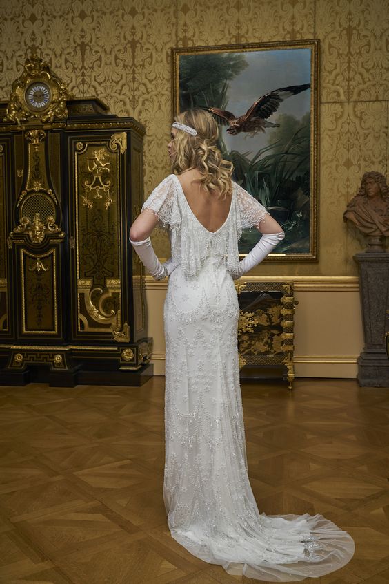 a 1920s inspired embellished lace wedding dress with a cutout back, wide sleeves, a train and tall gloves, an embellished headband