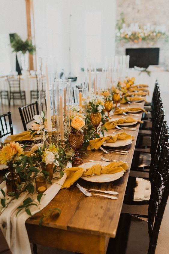a beautiful and bright fall wedding tablescape with greenery, mustard-colored blooms and napkins, tall candles, brown glassses and greenery