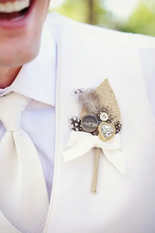 a beautiful wedding boutonniere of burlap, buttons, a heart rhinestone, feathers and a bow is a cool idea to make the groom's look bolder and catchier