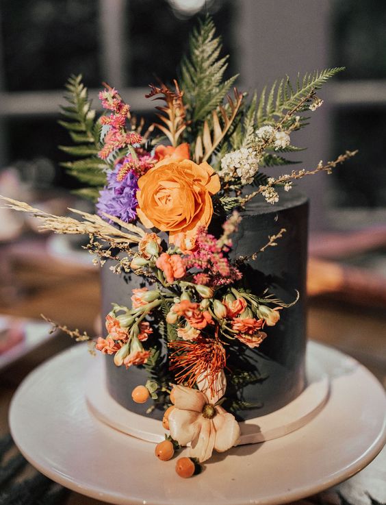 a black wedding cake decorated with fresh bold fall bloos and greenery and leaves
