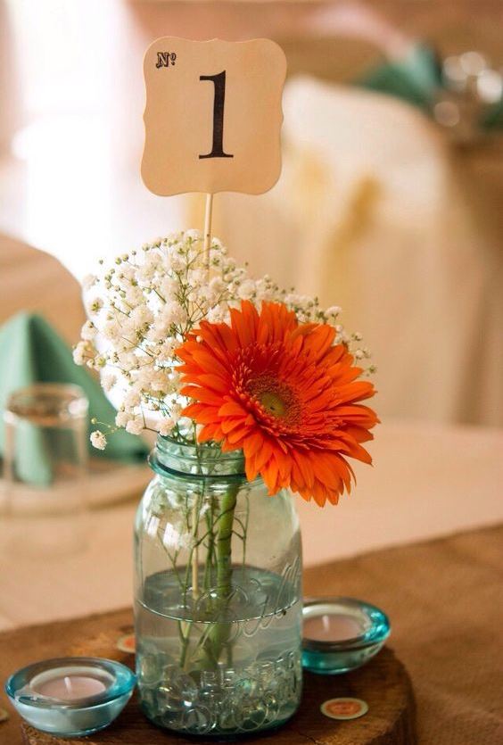 a blue jar with baby's breath and a bright dahlia plus a table number and candles make up a nice rustic centerpiece